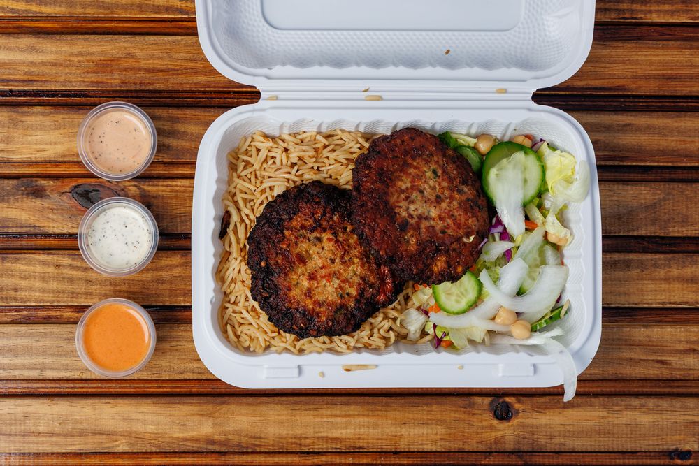 Two chapli kabab patties with dark, crisp edges, served over rice with a salad in a takeaway box beside three sauce cups.
